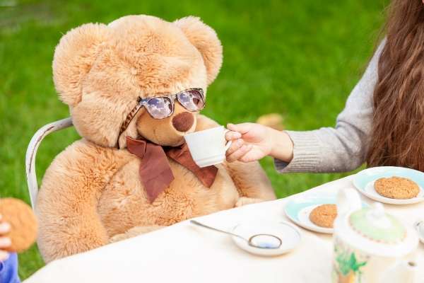 Parent, child and ted bear enjoying each a cup of tea and each a cookie.