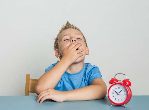 An image of a young boy yawning while seated with a clock on the table.